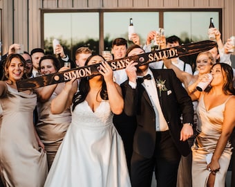 May include: A group of people celebrating, holding drinks and wooden signs that read "Simon Says Salud!" The bride wears a white wedding dress, and the groom wears a black tuxedo. The wedding party is dressed in formal attire, with a building and string lights in the background.