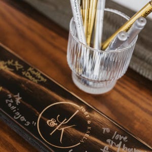 May include: A wooden sign with the names "Joey & Kaela" and well wishes written on it. A clear glass with gold and silver pens is next to the sign. The sign has a circular logo with the initials "J & K".