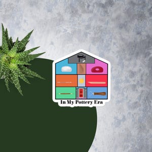 Puede incluir: Una pegatina con un diseño en forma de casa que presenta varias herramientas y elementos de cerámica en cuadrados de diferentes colores. El texto "In My Pottery Era" está en la parte inferior. La pegatina está sobre un fondo verde y gris.