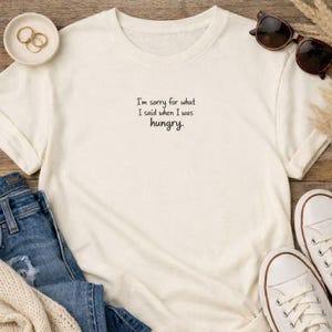 May include: Cream-colored t-shirt with the text "I'm sorry for what I said when I was hungry." The shirt is paired with blue jeans, white sneakers, and sunglasses. A small dish with gold rings is also visible.