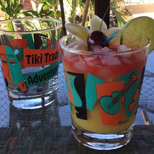 May include: Two clear Tiki Trade Adventure glasses, one filled with a colorful cocktail garnished with a cherry, lime, and flowers. The other glass is empty. The glasses have orange, teal, and black graphics.