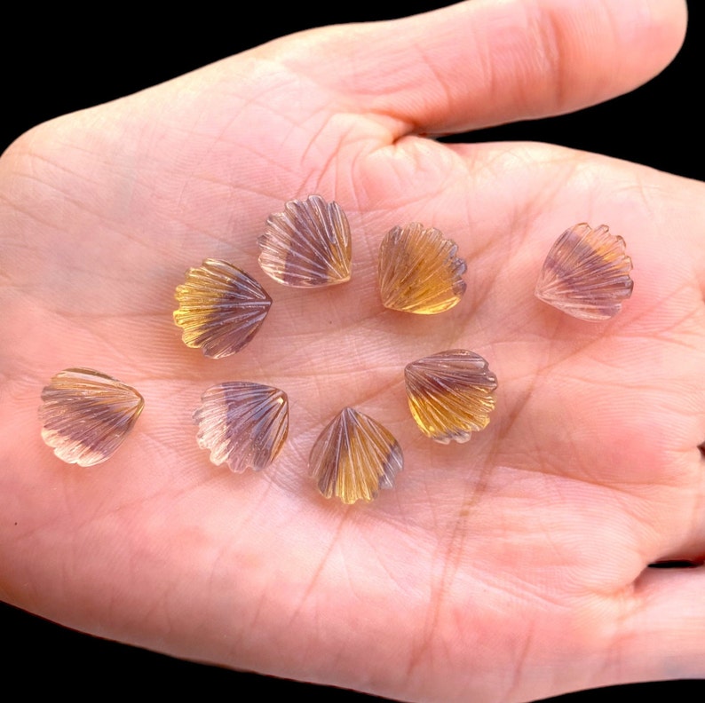 May include: A collection of eight iridescent, faceted, shell-shaped gemstone beads in shades of purple and yellow. The beads are arranged on a hand.