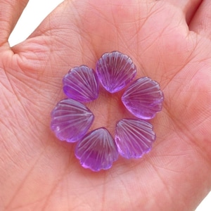 May include: Six purple amethyst gemstone cabochons shaped like seashells. The cabochons are arranged in a circle on a hand.