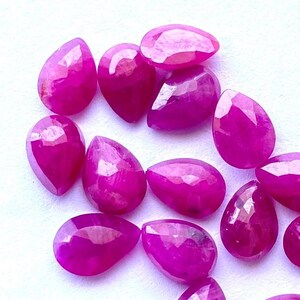 Puede incluir: Una colecci&oacute;n de gemas facetadas en forma de l&aacute;grima, de un vibrante color magenta. Las gemas est&aacute;n dispersas sobre una superficie blanca, mostrando sus superficies pulidas y su claridad interna. Ideal para la fabricaci&oacute;n de joyas o coleccionar.