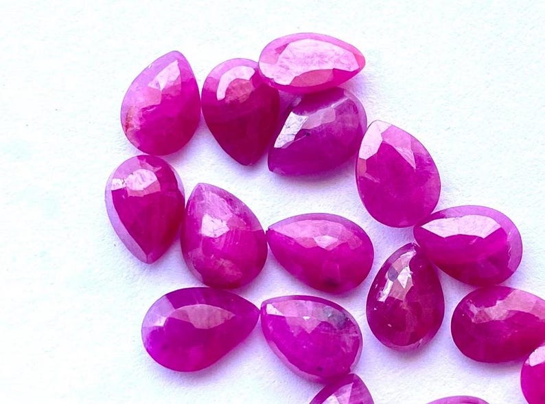 Puede incluir: Una colecci&oacute;n de gemas facetadas en forma de l&aacute;grima, de un vibrante color magenta. Las gemas est&aacute;n dispersas sobre una superficie blanca, mostrando sus superficies pulidas y su claridad interna. Ideal para la fabricaci&oacute;n de joyas o coleccionar.