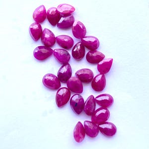 Puede incluir: Un conjunto de gemas facetadas en forma de l&aacute;grima, de un vibrante color magenta. Las gemas est&aacute;n dispersas sobre una superficie blanca, mostrando sus superficies pulidas y claridad interna. Adecuadas para la creaci&oacute;n de joyas. Medidas en cent&iacute;metros.