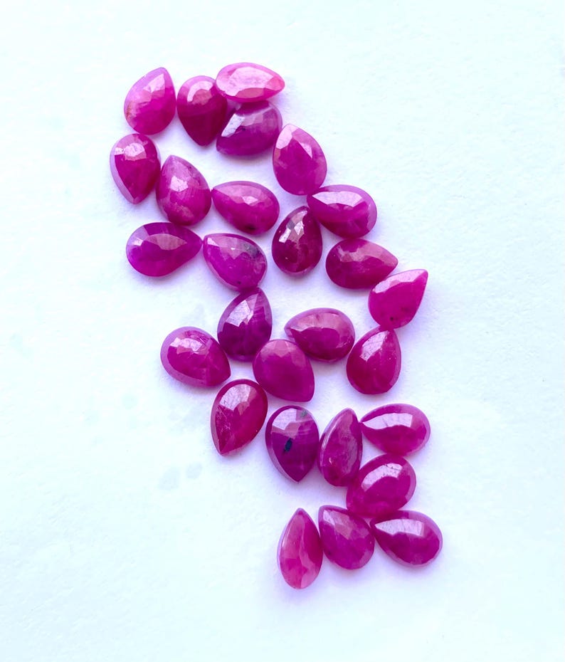 Puede incluir: Un conjunto de gemas facetadas en forma de l&aacute;grima, de un vibrante color magenta. Las gemas est&aacute;n dispersas sobre una superficie blanca, mostrando sus superficies pulidas y claridad interna. Adecuadas para la creaci&oacute;n de joyas. Medidas en cent&iacute;metros.