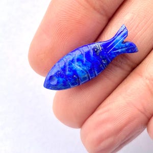 Puede incluir: Pequeña figura de pez tallada en lapislázuli. El pez es de color azul oscuro con vetas azules y doradas más claras. La cola y las aletas están detalladas. La figura se sostiene en una mano.