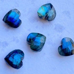 Puede incluir: Cinco gemas de labradorita en forma de corazón con superficies facetadas. Las piedras muestran un vibrante juego de colores, principalmente azules y verdes, con toques dorados. Cada corazón mide aproximadamente 2.5 cm de ancho.