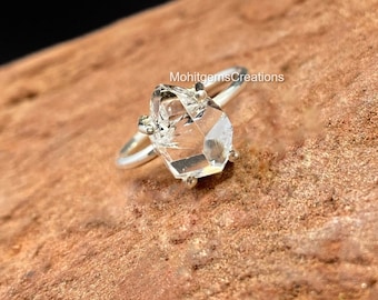 May include: A silver ring with a clear, faceted, Herkimer diamond. The diamond is set in a bezel setting with four prongs. The ring is on a brown wooden surface.