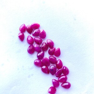 Puede incluir: Colecci&oacute;n de rub&iacute;es facetados en forma de l&aacute;grima, de varios tama&ntilde;os. Las piedras preciosas son de un color rojo rosado intenso y vibrante, dispersas sobre una superficie blanca. Las gemas tienen una superficie pulida y reflectante.