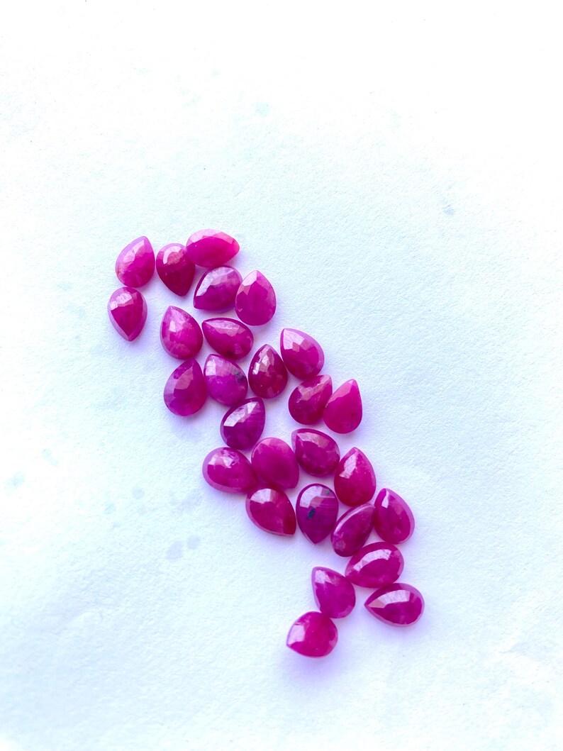 Puede incluir: Colecci&oacute;n de rub&iacute;es facetados en forma de l&aacute;grima, de varios tama&ntilde;os. Las piedras preciosas son de un color rojo rosado intenso y vibrante, dispersas sobre una superficie blanca. Las gemas tienen una superficie pulida y reflectante.