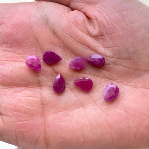 Puede incluir: Colecci&oacute;n de siete gemas de rub&iacute; facetadas, en forma de l&aacute;grima y ovalada. Los rub&iacute;es var&iacute;an en tama&ntilde;o y tienen un color rosa intenso a magenta. Las gemas se muestran sobre un fondo de tono de piel claro.