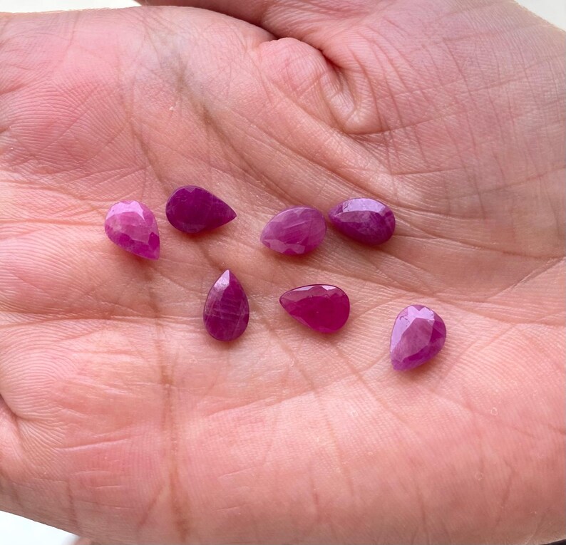 Puede incluir: Colecci&oacute;n de siete gemas de rub&iacute; facetadas, en forma de l&aacute;grima y ovalada. Los rub&iacute;es var&iacute;an en tama&ntilde;o y tienen un color rosa intenso a magenta. Las gemas se muestran sobre un fondo de tono de piel claro.