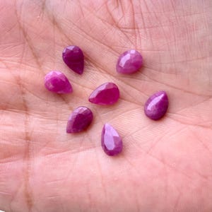 Puede incluir: Siete gemas de rub&iacute; facetadas en forma de l&aacute;grima, en varios tonos de rosa y rojo, se muestran en una mano. Las gemas tienen un aspecto pulido y brillante y est&aacute;n dispuestas en un patr&oacute;n disperso.