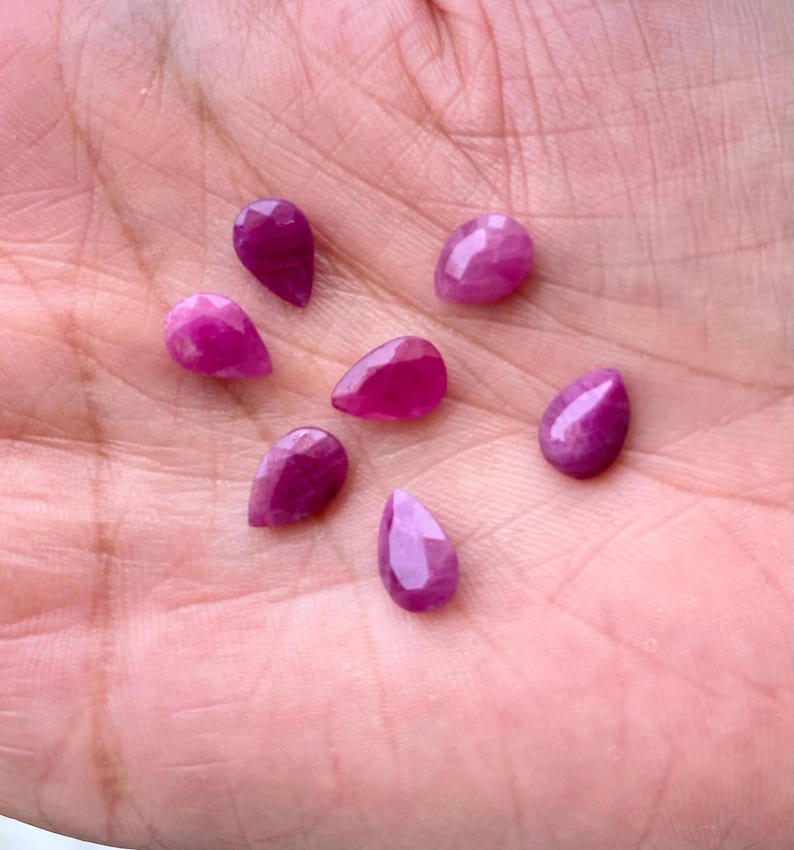 Puede incluir: Siete gemas de rub&iacute; facetadas en forma de l&aacute;grima, en varios tonos de rosa y rojo, se muestran en una mano. Las gemas tienen un aspecto pulido y brillante y est&aacute;n dispuestas en un patr&oacute;n disperso.