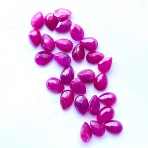 May include: A collection of faceted, teardrop-shaped, deep pink gemstones. The gems are scattered on a white surface, each displaying a polished, reflective surface. The arrangement highlights the individual stones' color and cut, suitable for jewelry making.