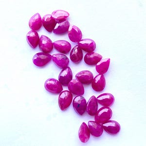 Puede incluir: Colecci&oacute;n de gemas de rub&iacute; pulidas en forma de l&aacute;grima. Las gemas son de un color magenta vibrante y est&aacute;n dispersas sobre una superficie blanca. Cada piedra tiene una superficie facetada, que atrapa la luz y crea un efecto brillante. Ideal para la fabricaci&oacute;n de joyas.