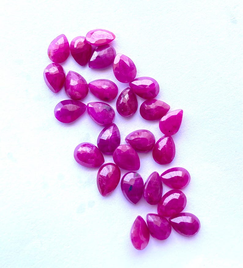 Puede incluir: Colecci&oacute;n de gemas de rub&iacute; pulidas en forma de l&aacute;grima. Las gemas son de un color magenta vibrante y est&aacute;n dispersas sobre una superficie blanca. Cada piedra tiene una superficie facetada, que atrapa la luz y crea un efecto brillante. Ideal para la fabricaci&oacute;n de joyas.