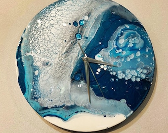 Puede incluir: Un reloj redondo con un diseño abstracto azul y blanco. La esfera del reloj está hecha de resina y tiene una aguja de reloj de metal plateado.