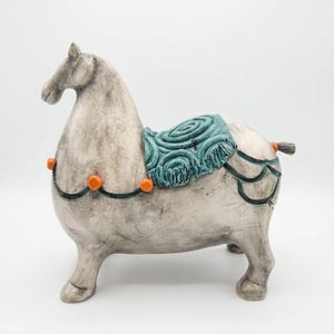Puede incluir: Figura de caballo de cerámica en color gris claro con una silla de montar turquesa y detalles en naranja. El caballo tiene un diseño detallado y una estética decorativa. La figura es un objeto decorativo.