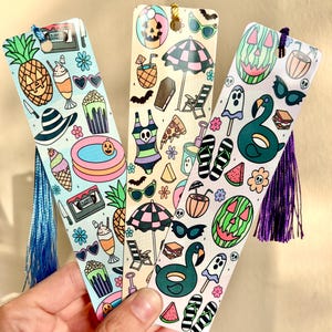 May include: Three colorful bookmarks with summer and Halloween-themed illustrations. One features a pineapple, ice cream, and a pool. Another has a beach umbrella, coffin, and pizza. The last one has a watermelon, ghost, and a swan.