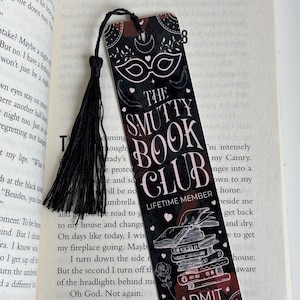 May include: A black and pink bookmark with a black tassel. The bookmark features a masquerade mask, a stack of books, and the text "The Smutty Book Club - Lifetime Member" and "Admit One".