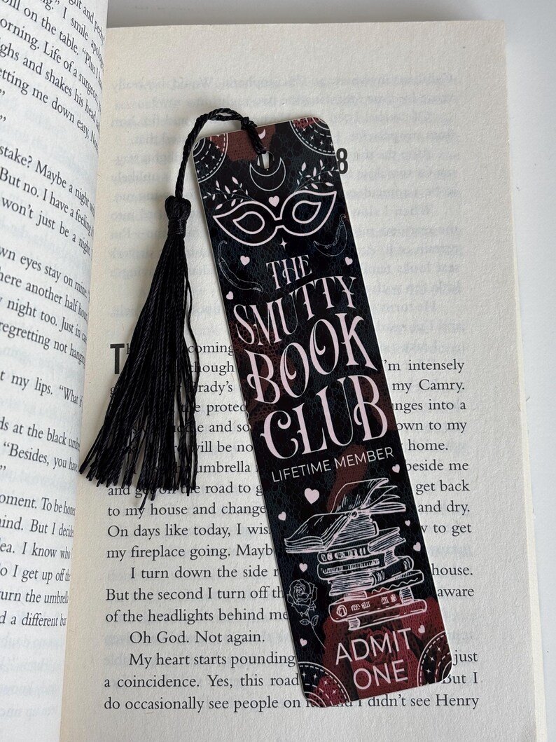 May include: A black and pink bookmark with a black tassel. The bookmark features a masquerade mask, a stack of books, and the text "The Smutty Book Club - Lifetime Member" and "Admit One".