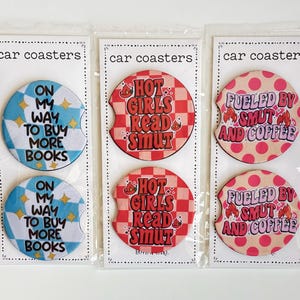 May include: Three sets of car coasters in clear packaging. One set has blue and white checkered coasters with the text "ON MY WAY TO BUY MORE BOOKS." Another set has red checkered coasters with the text "HOT GIRLS READ SMUT." The last set has pink polka dot coasters with the text "FUELED BY SMUT AND COFFEE."
