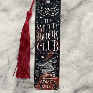 May include: A black and red bookmark with a masquerade mask, the text "The Smutty Book Club", and "Lifetime Member" printed on it. The bookmark also features a stack of books, a rose, and the text "Admit One".