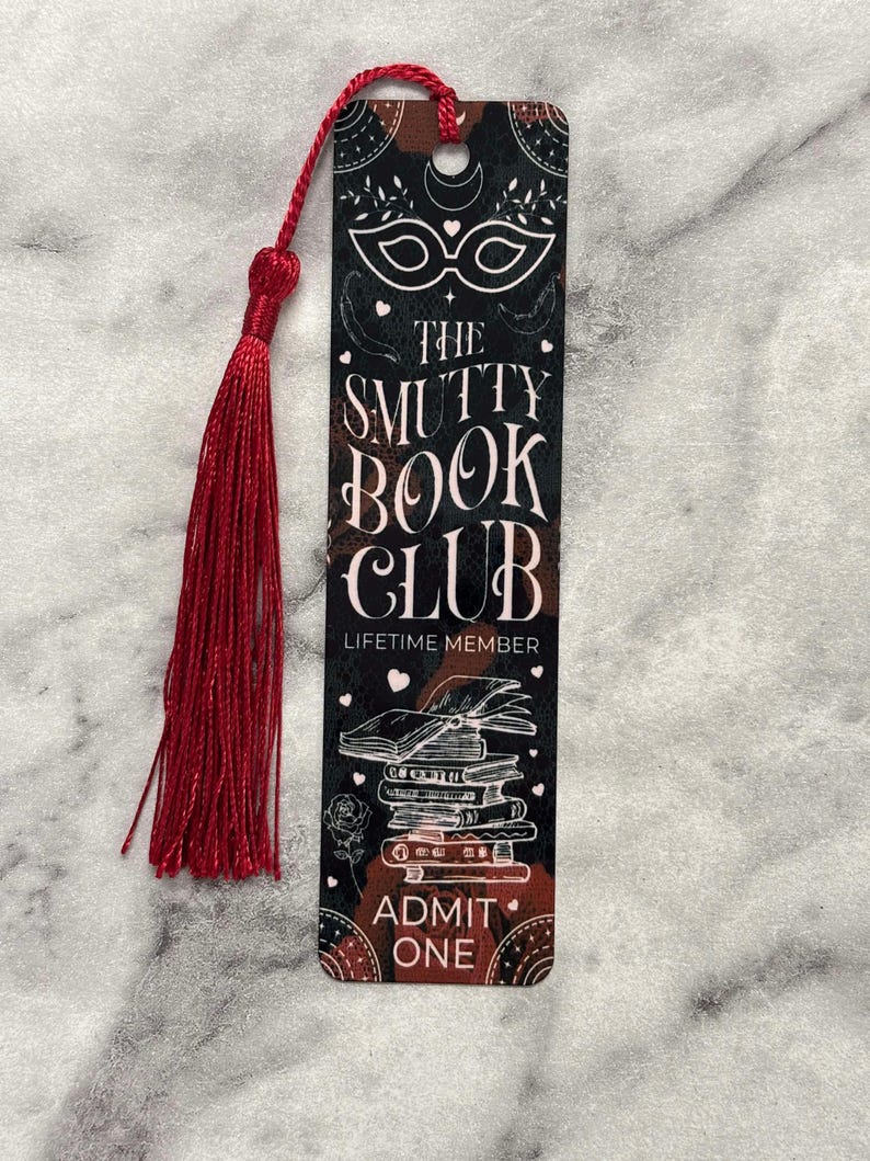 May include: A black and red bookmark with a masquerade mask, the text "The Smutty Book Club", and "Lifetime Member" printed on it. The bookmark also features a stack of books, a rose, and the text "Admit One".