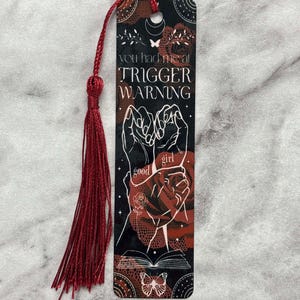 May include: A black and red bookmark with a white design. The bookmark features two hands holding each other, a butterfly, and the text "You had me at trigger warning".