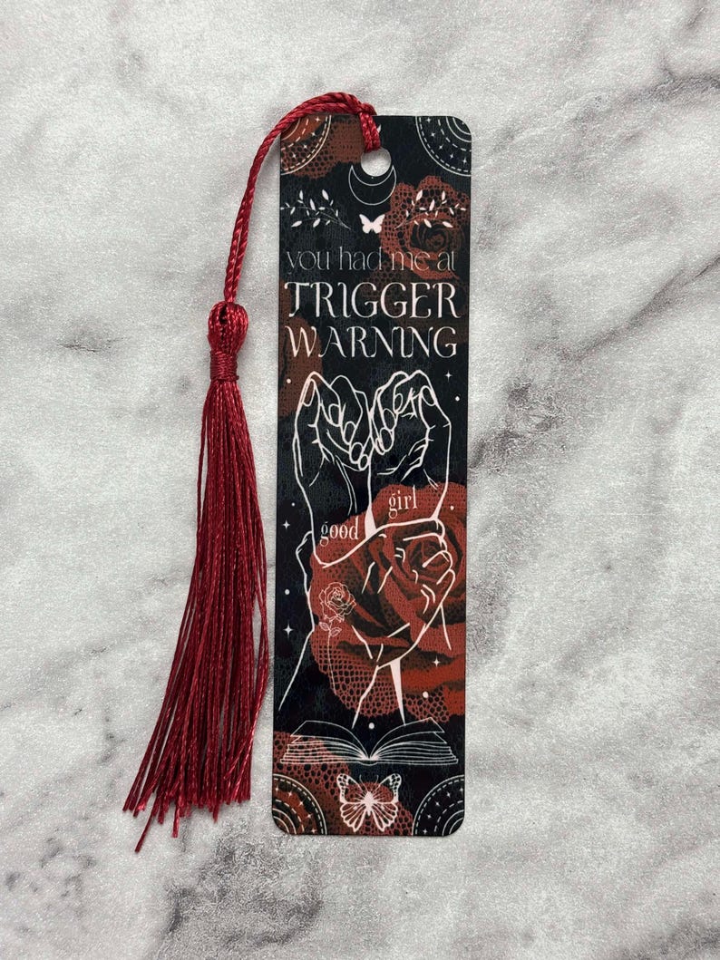 May include: A black and red bookmark with a white design. The bookmark features two hands holding each other, a butterfly, and the text "You had me at trigger warning".