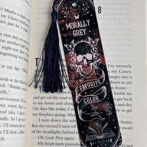 May include: A black and red bookmark with a skull and crossbones design. The bookmark has a tassel and the text "Morally Grey. Is My Favorite Color."