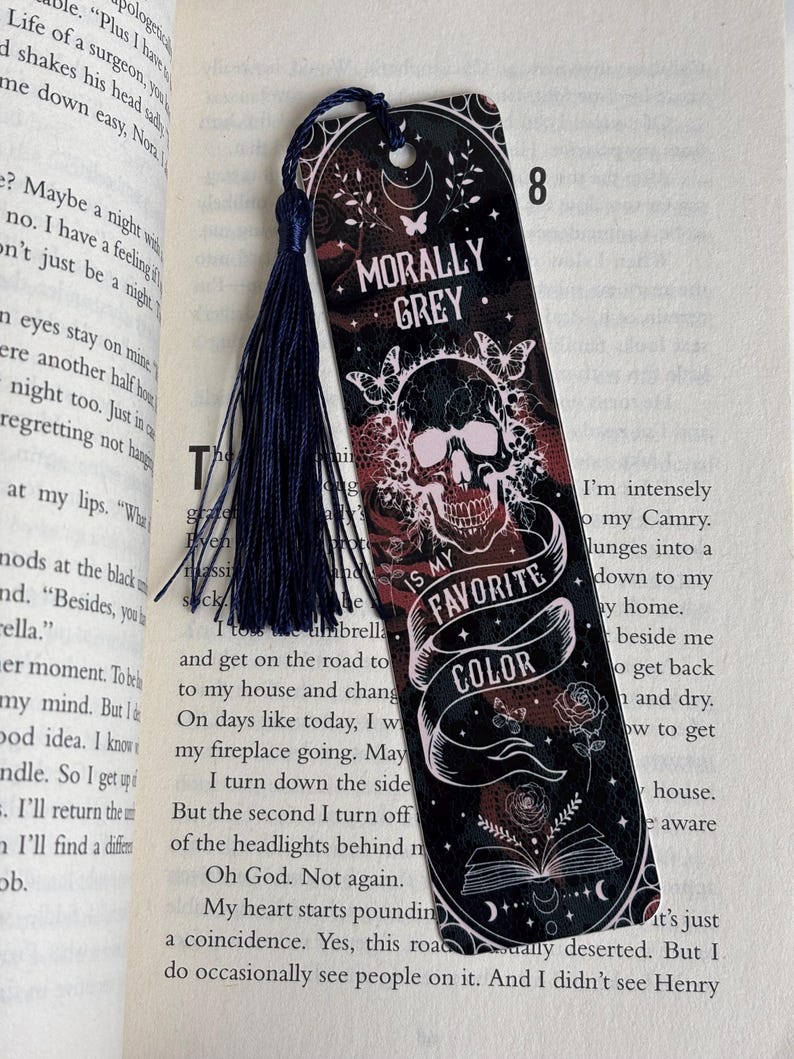 May include: A black and red bookmark with a skull and crossbones design. The bookmark has a tassel and the text "Morally Grey. Is My Favorite Color."