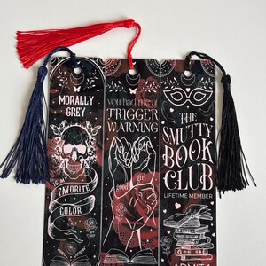 May include: Set of three black and red bookmarks with a skull, hands, and a mask. The bookmarks have white text that reads "Morally Grey is my favorite color", "You had me at Trigger Warning", and "The Smutty Book Club Lifetime Member Admit One".