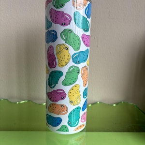 May include: A white stainless steel tumbler with a colorful pattern of cartoon clogs. The clogs are in various colors, including green, yellow, blue, pink, and orange.