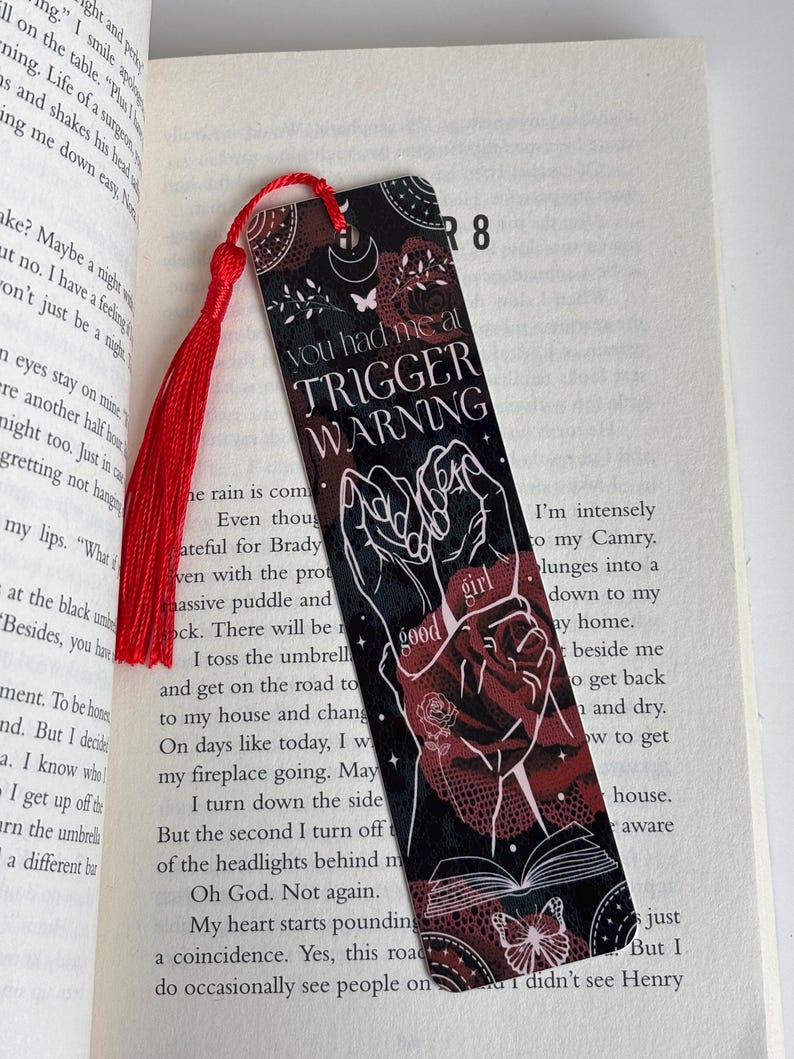 May include: A black and red bookmark with a white butterfly and the text "You had me at Trigger Warning". The bookmark features a stylized image of two hands clasped together with a red rose and the words "good girl" in white.