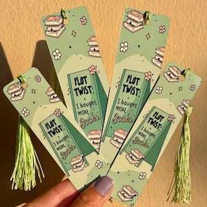 May include: Four green and white bookmarks with a light green tassel. Each bookmark has the text "PLOT TWIST: I bought MORE Books" with illustrations of books and flowers. The bookmarks are held in a hand.