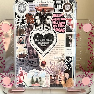 May include: A clear Kindle case decorated with stickers featuring text and images. The stickers include a heart with the text "This is the Kindle of a reader, Bella", images of a red truck, and other graphics. The case is displayed on a pink stand.