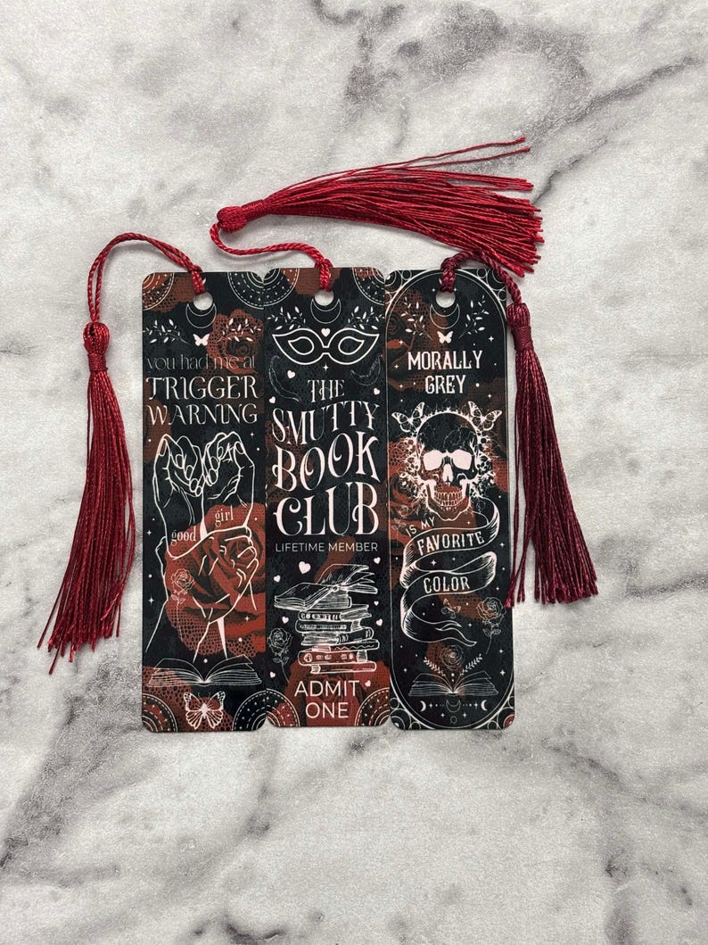 May include: Three bookmarks with red tassels. The bookmarks feature a black and red background with white text and illustrations. The text on the bookmarks reads "You had me at trigger warning", "The Smutty Book Club", and "Morally Grey is my favorite color".