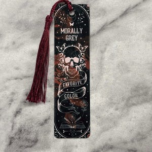 May include: A black and red bookmark with a skull and crossbones design. The bookmark has a tassel and the text "Morally Grey is my favorite color".
