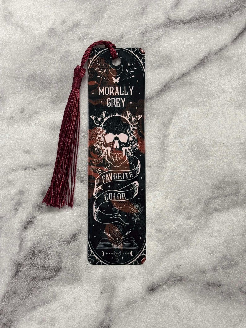 May include: A black and red bookmark with a skull and crossbones design. The bookmark has a tassel and the text "Morally Grey is my favorite color".