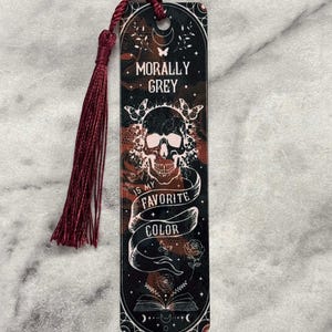 May include: A black and red bookmark with a skull and the text "Morally Grey is my favorite color". The bookmark has a red tassel.