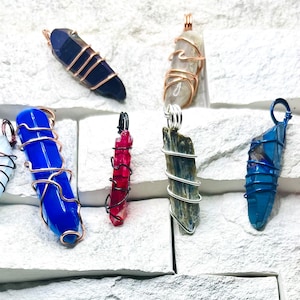 Può includere: Una collezione di 11 ciondoli di cristallo avvolti in filo di rame. I cristalli sono di vari colori, tra cui verde, blu, rosa, rosso e trasparente. I ciondoli sono tutti lunghi circa 5 cm.