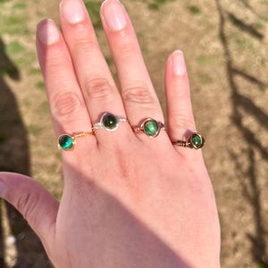 May include: Four rings with green gemstone settings. The rings are made of gold wire wrapped around the stones. The stones are round and have a dark green color.