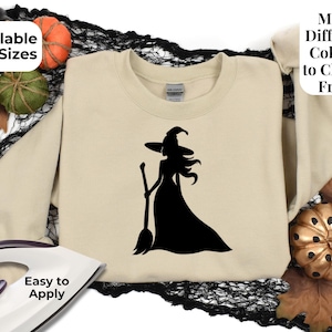 May include: A beige crewneck sweatshirt featuring a black silhouette graphic of a witch wearing a pointed hat and holding a broom. The image includes text that reads Available in 3 Sizes, Many Different Colours to Choose From, and Easy to Apply.