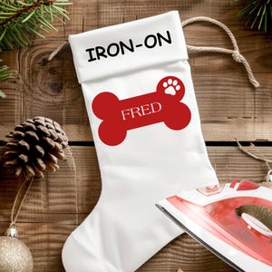 May include: White Christmas stocking with the text "IRON-ON" at the top. A red bone shape with the name "FRED" and a paw print is printed on the stocking. A red and white iron is in the bottom right corner.