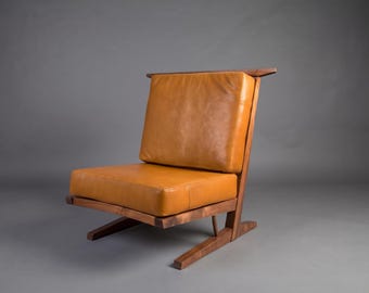 May include: A low-profile lounge chair featuring a dark brown wooden frame with angled legs and a horizontal back support. The seat and backrest consist of two thick, rectangular cushions upholstered in smooth, tan-colored leather.