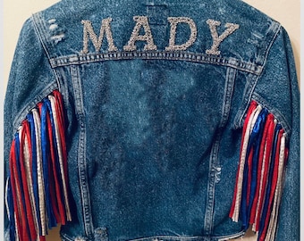 May include: A blue denim jacket with red, white, and blue fringe on the sleeves. The back of the jacket has the name "MADY" spelled out in silver rhinestones.